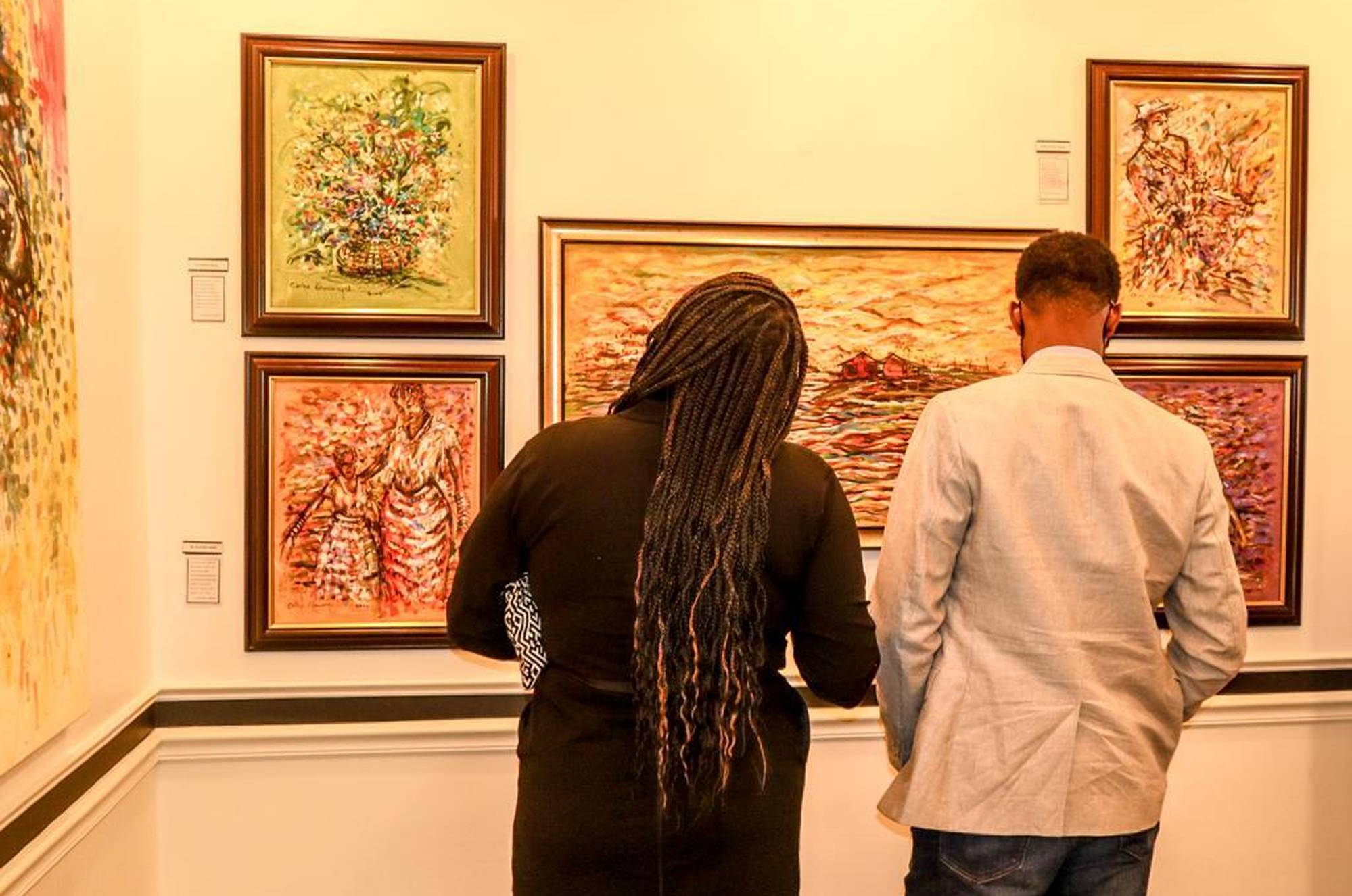 A woman and man viewing a collection of framed African paintings in a warmly lit gallery. The artworks feature expressive brushstrokes, vibrant colors, and emotionally rich scenes, reflecting rare themes and deep cultural meaning—similar to the pieces Cheryl discovered at Ovico Gallery.